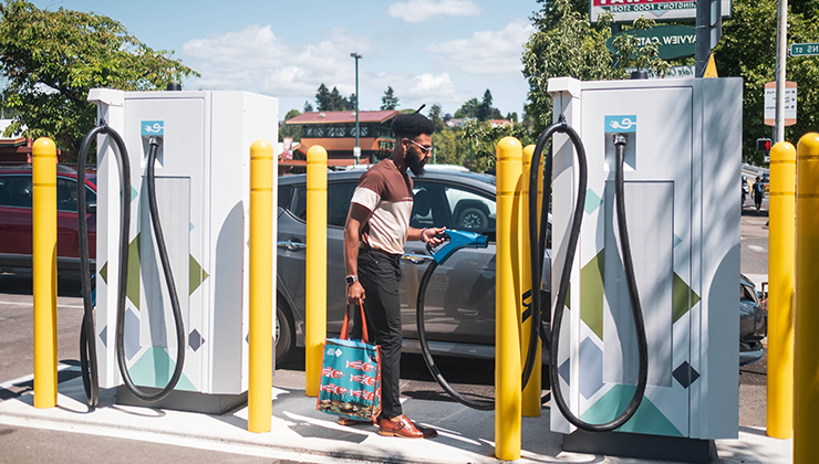 A man with a shopping bag in one hand and an EV charging nozzle in the other hand, after charging his car at an EV charging station